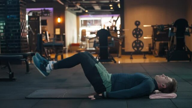 Athletic man doing leg raises lying on yoga mat, focused on abdominal muscle toning, within the vibrant dim ambiance of a modern gym - profile view