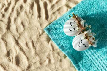 Two chicken eggs with hand drawn happy faces with wreaths lie on a beach towel on the sand. Shadow from palm leaves.