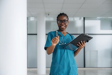 In glasses, standing. Female doctor is indoors in modern room