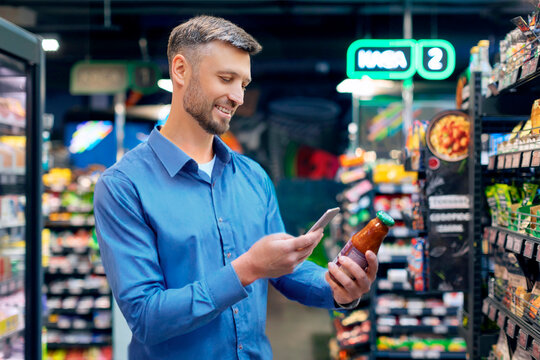 Smiling man scanning qr code on glass jar with smartphone, supermarket male customer using an online application and scanning barcode of products