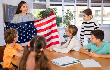 Group of curious preteen learners and young female teacher with flag of USA in classroom of academy