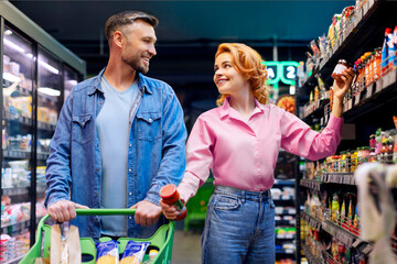 Obraz premium Happy spouses shopping together, buying food at supermarket, walking with shopping cart choosing products and talking