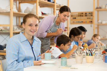 Positive teenage girl painting handmade ceramic cup with brush in pottery workshop