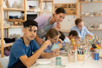 Smiling teen boy coloring handmade clay jar with paintbrush in art studio