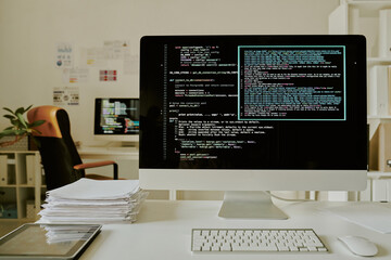 Medium close up of computer with code lines on monitor, stack of documents and tablet on top of desk in office