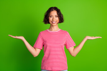 Smiling young woman presenting with open hands in casual attire against a vibrant green background in a stylish striped t-shirt