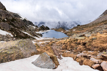 Mountain lake surrounded by rocks and snow, with clouds in the background. Serene high-altitude lake surrounded by rocky, snowy terrain under a cloudy sky. GR11, Lleida