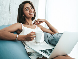 Obraz premium Portrait of beautiful smiling woman dressed in white pajamas. Carefree model sitting on soft bag chair and using laptop. Sexy female reading news and enjoying her morning at balcony, drinking coffee
