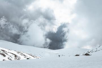 Hiker crossing a snowy alpine valley beneath dramatic storm clouds in Sierra de Gredos, Spain,...