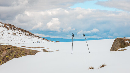 Abandoned trekking poles in snowy Sierra de Gredos, Spain, with a group of hikers in the distance and a dramatic sky above a remote winter mountain landscape