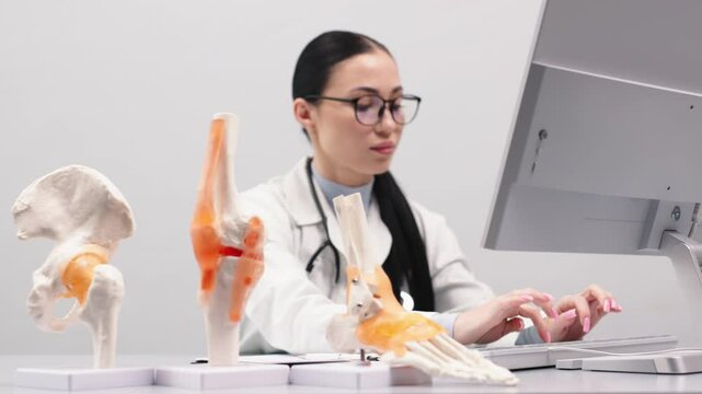 An orthopedic specialist works in a modern office using a computer. Models of the knee joint, pelvic floor and foot are on the table. Concept of orthopedic diagnostics.