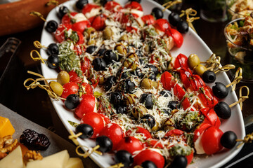 Skewers of vibrant caprese salad featuring cherry tomatoes, fresh mozzarella, black and green olives, drizzled with pesto sauce and sprinkled with grated cheese on a platter