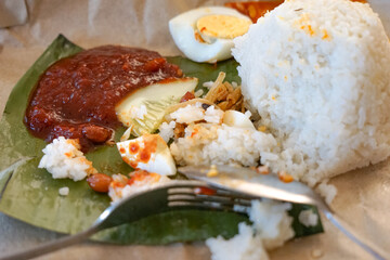 A serving of Nasi Lemak featuring rice cooked with coconut milk and herbs, hard-boiled egg with spicy sambal, fresh cucumber slices, and crispy fried anchovies on a banana leaf over brown paper.