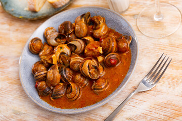 Stewed snails in red sauce served on a gray ceramic plate close to the cutlery and bread in the restaurant