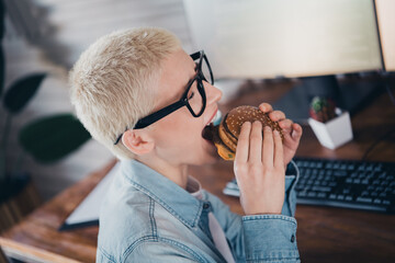 Young woman in denim shirt enjoying a burger while working on a computer in a cozy home office