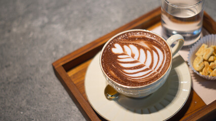 Close-up top view of coffee in a white glass ceramics decorated with heart pattern served with letter cookies and water on a wooden tray, creating a warm atmosphere and leaving space for text.