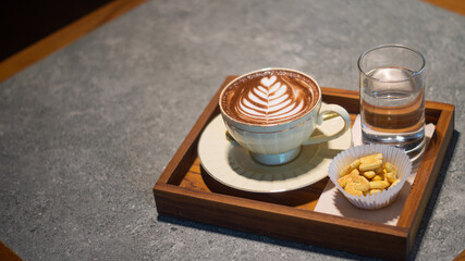 Coffee in a white glass ceramics decorated with heart pattern served with letter cookies and water placed on a warm wooden tray with space for text.