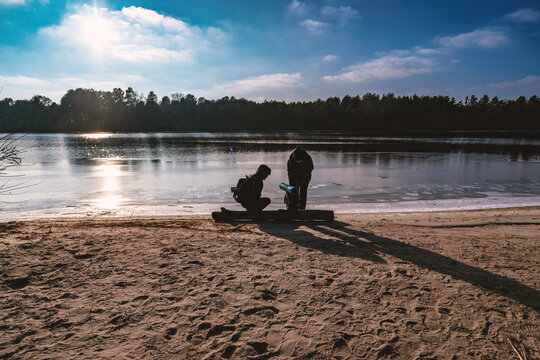 Two people pause by a frozen lake at sunset, casting long shadows on the sand as they prepare for a winter adventure. A peaceful, reflective moment in nature's cold beauty.