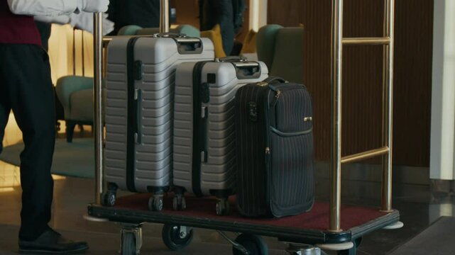 Close up view of bellhop in formal uniform placing suitcases on luggage cart in hotel lobby, then rolling it away