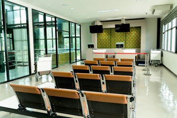 Empty airport boarding gate area with rows of brown waiting chairs, modern check-in counters, digital screens, and trolley carts in bright terminal space