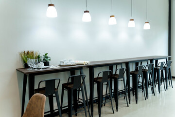 Modern cafe bar interior with high wooden counter, metal bar stools, wine glasses, tea set, pendant lights, and minimal white wall background