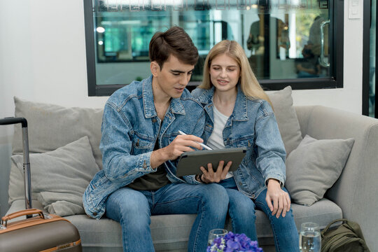 Young couple using digital tablet together on sofa in modern lounge, planning travel itinerary or working remotely while waiting with luggage - Powered by Adobe