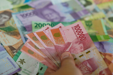 Hands holding several hundred thousand rupiah bills with a background of various types of Indonesian money. Suitable for background, finance, banking, cash, savings.