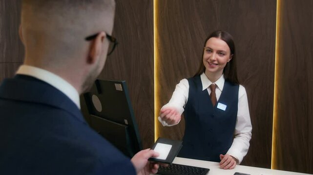 Female hotel receptionist typing guest information on computer, then giving him room key folder with passport while assisting with check-in at front desk