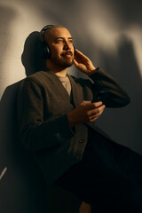 A caucasian man with a beard wearing a cardigan sitting alone using a cellphone and a pair of wireless headphones listening. 