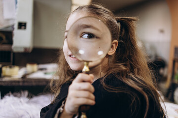 A young girl with long hair holds a magnifying glass, smiling confidently. She is in a kitchen...