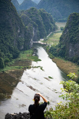 Naklejka premium (Selective focus) Defocused girl taking pictures from the Hang Múa Viewpoint with the a stunning landscape and the Tam Coc river in the distance. Ninh Binh, Vietnam.