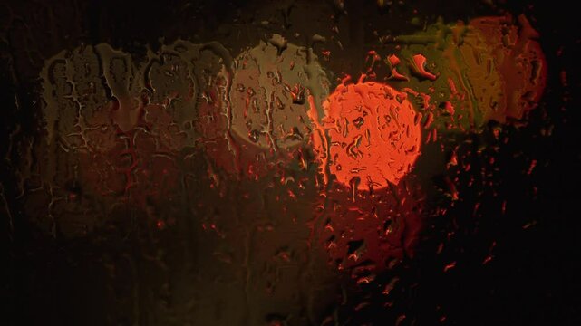 Raindrops in focus on a window, the darkness of the night is broken by trafic lights and the glow of car headlights. Atmospheric and cinematic.