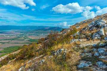 Panoramic view from a Crimean slope to a valley and a village below. In the foreground is a rocky mountain path covered with dry grass and bushes. 