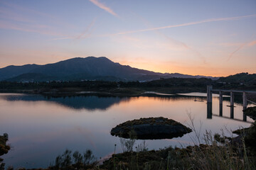 Calm lake reflecting mountain range at sunset with concrete bridge structure