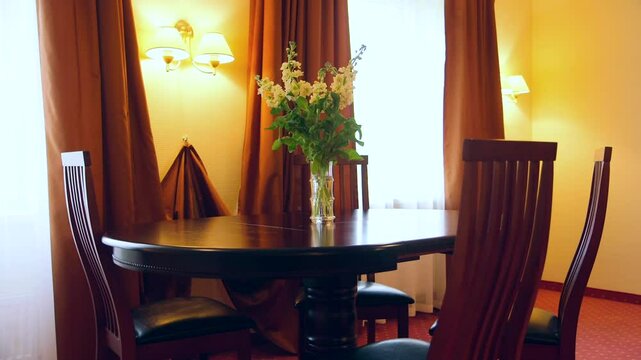 In the Hotel Room, there is an Elegant Wooden Table with Four Chairs. On the Table are White Flowers in a Vase. Red Carpet. Two Windows with Half-Open Curtains. Two Lit Wall Lamps.