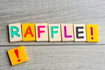 Colorful wooden letter tiles, spelling "RAFFLE", exclamation mark, light gray wooden background, vibrant primary colors, close-up photography, game pieces, word formation, fundraising concept