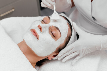 Woman getting professional skin facial care treatment procedures at spa and beauty salon. Young lady with white towel turban on head lying on bed while beautician applies cleansing mask on her face