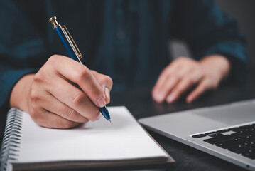 Close-up hand writing on notebook with pen next to laptop. Education, business planning, journaling setup with modern workspace productivity and creative thinking.