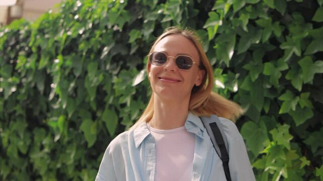 Blonde tourist woman wearing sunglasses,  light blue shirt and  t-shirt smiles and touche hair and sunglasses in front of green ivy wall.Europe travel.Lifestyle,vacation,tourism,nature,active life