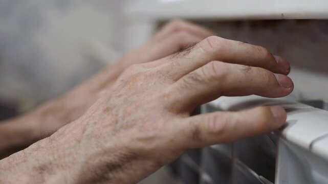 Close-up of elderly hands on a radiator capturing the difficulty of staying warm at home during the cold season, emphasizing the impact of energy shortages and the struggle for basic comfort