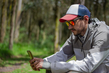 A person in a gray and white waterproof jacket with a red-brim cap holds a smartphone amid lush...