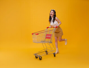 Full length happy excited Asian teen woman pushing empty shopping cart or shopping trolley grocery running promotion supermarket isolated on yellow background. © NaMong Productions
