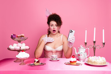 Young woman in elegant corset and gloves holds smartphone at an elaborate pink afternoon tea table
