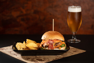 Tasty hamburger with dried bacon, cheese and chips served in a black plate with a glass of beer on dark ambient background, stock