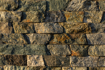 Rough stacked stone wall texture with brown and gray tones, showing layered rocks with visible shadows and detail.
