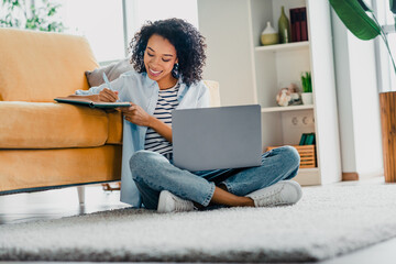 Young woman working and relaxing on the floor of a cozy living room, writing notes and using a...