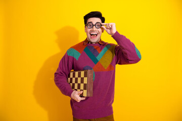 Young man with glasses holding a chessboard in a colorful sweater against yellow background