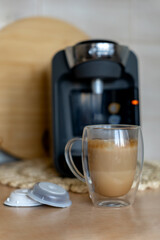 Cup of latte with foam and capsules for brewing coffee on the background of capsule coffee maker in home kitchen.