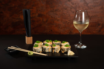 still life of black plate with fried uramaki, ginger  nd wasabi on dark background, with white wine and chopsticks
