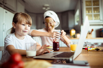 Single mother and daughter having breakfast together in the morning while using a laptop in the kitchen